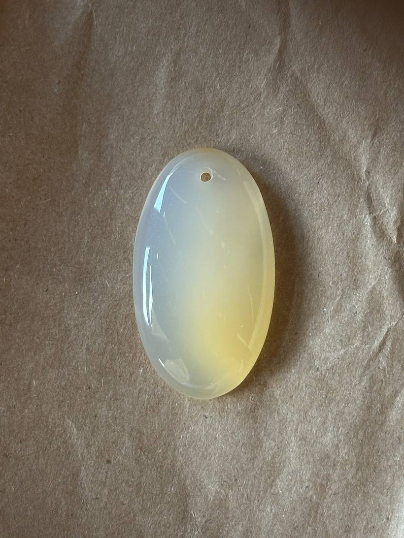 An oval-shaped translucent pendant made of milky white stone with a soft yellow tint, resting on a textured brown paper background.