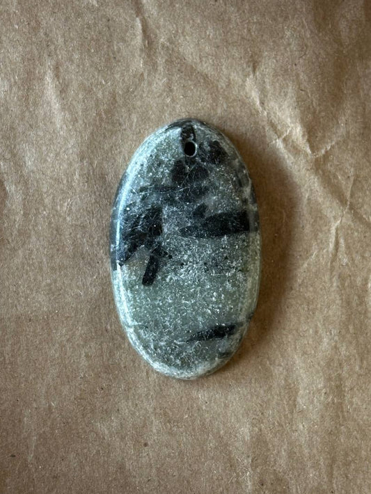 An oval stone pendant with a natural marbled pattern of black and gray, featuring a small hole at the top for hanging, displayed on a brown textured paper background.