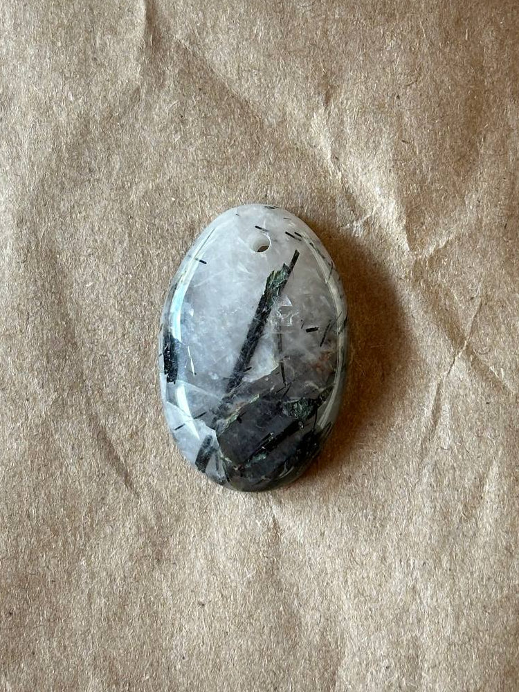 A polished oval gemstone with a white and gray base and dark green and black inclusions, resting on a textured brown kraft paper background.
