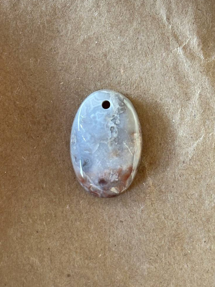 An oval polished stone pendant with a natural marbled pattern in gray, white, and brown, featuring a small hole at the top, resting on a textured brown paper background.