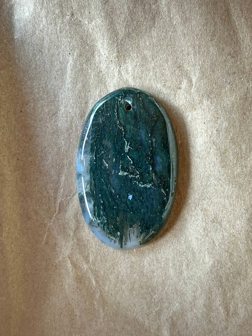 An oval polished stone pendant with a dark teal and green marbled pattern, featuring a small hole at the top for hanging, displayed on a textured beige paper background.