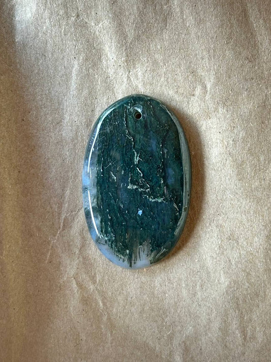 An oval polished stone pendant with a dark teal and green marbled pattern, featuring a small hole at the top for hanging, displayed on a textured beige paper background.