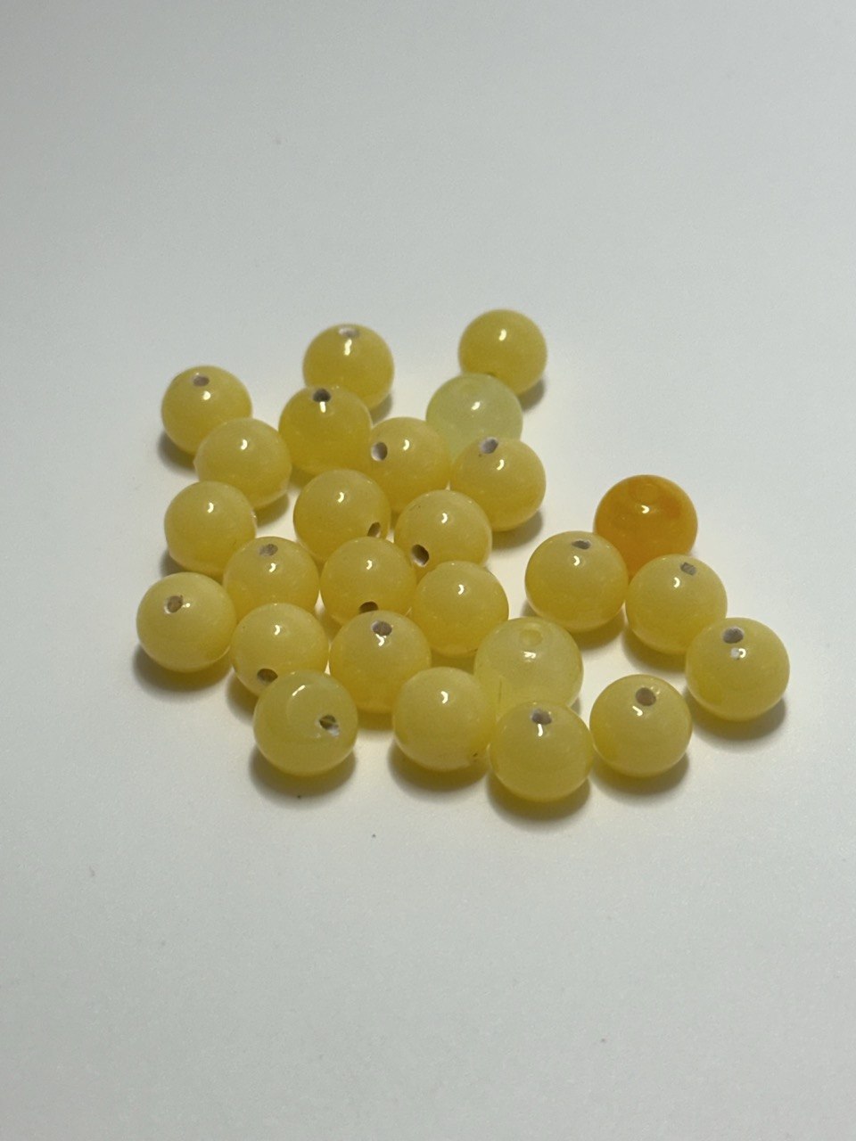 A pile of small round glass yellow beads with holes, photographed on a plain white background. One bead near the upper right is a slightly deeper amber-orange tone.