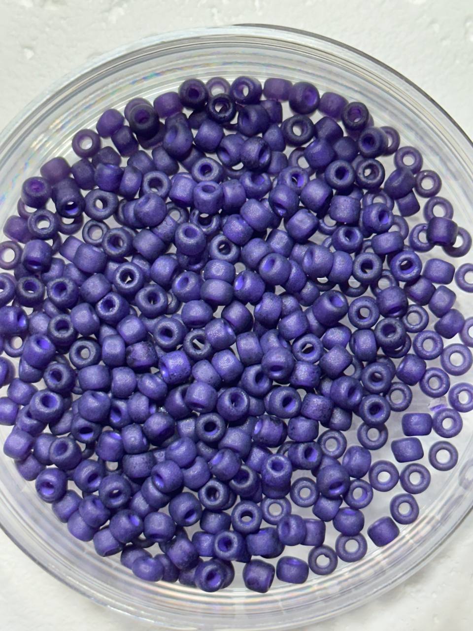 A top-down view of small, cylindrical purple seed beads with a metallic sheen, piled inside a clear glass bowl on a light textured background.