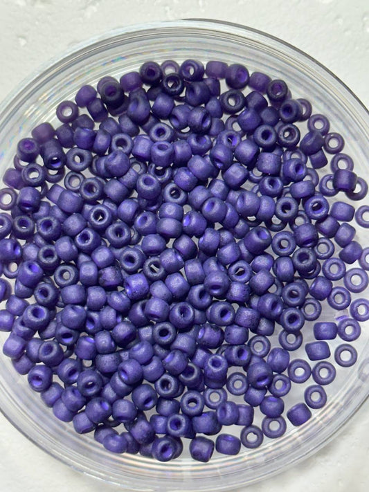 A top-down view of small, cylindrical purple seed beads with a metallic sheen, piled inside a clear glass bowl on a light textured background.