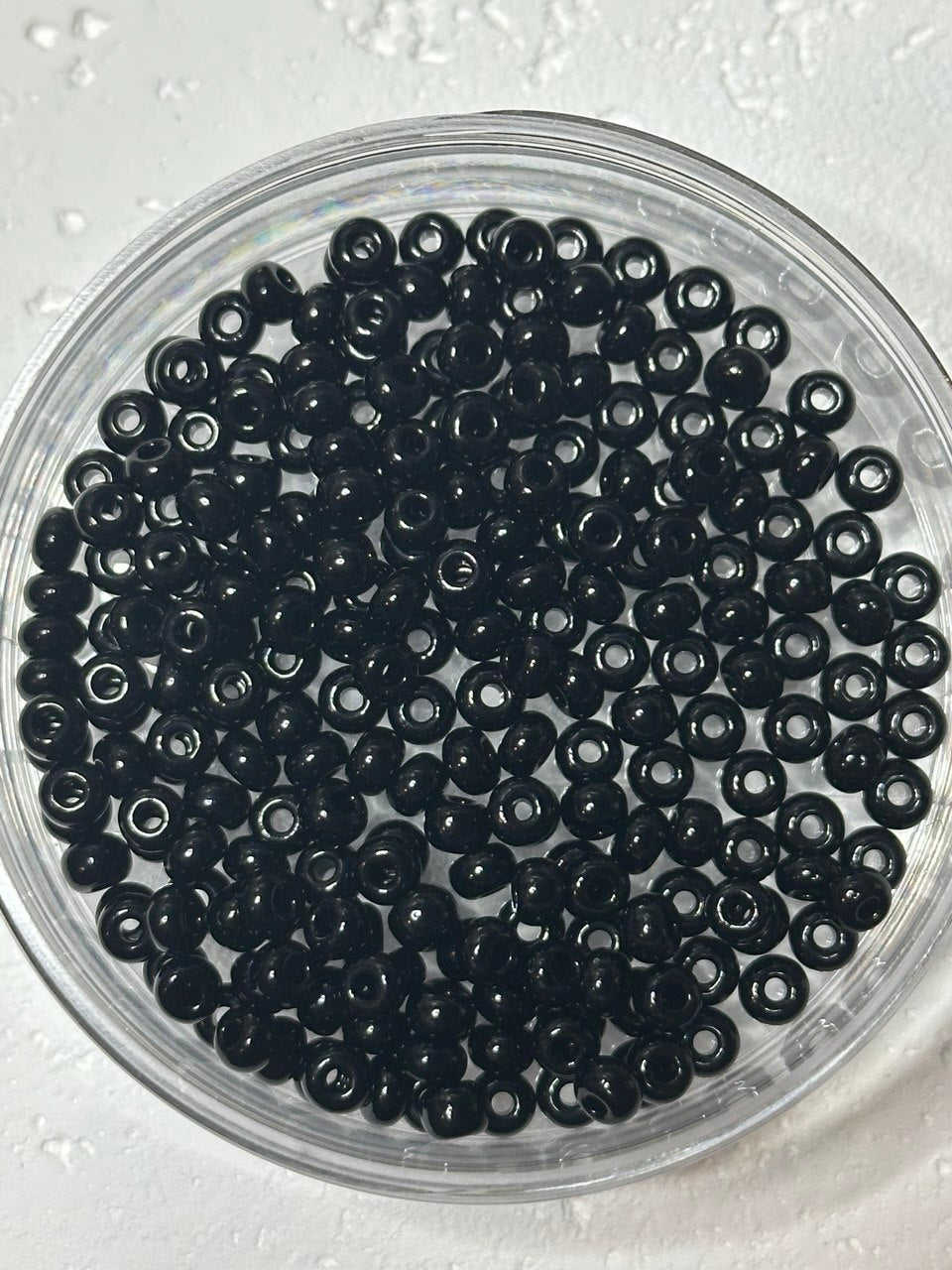 Black seed beads with a glossy finish, arranged inside a clear plastic round container on a light textured background.