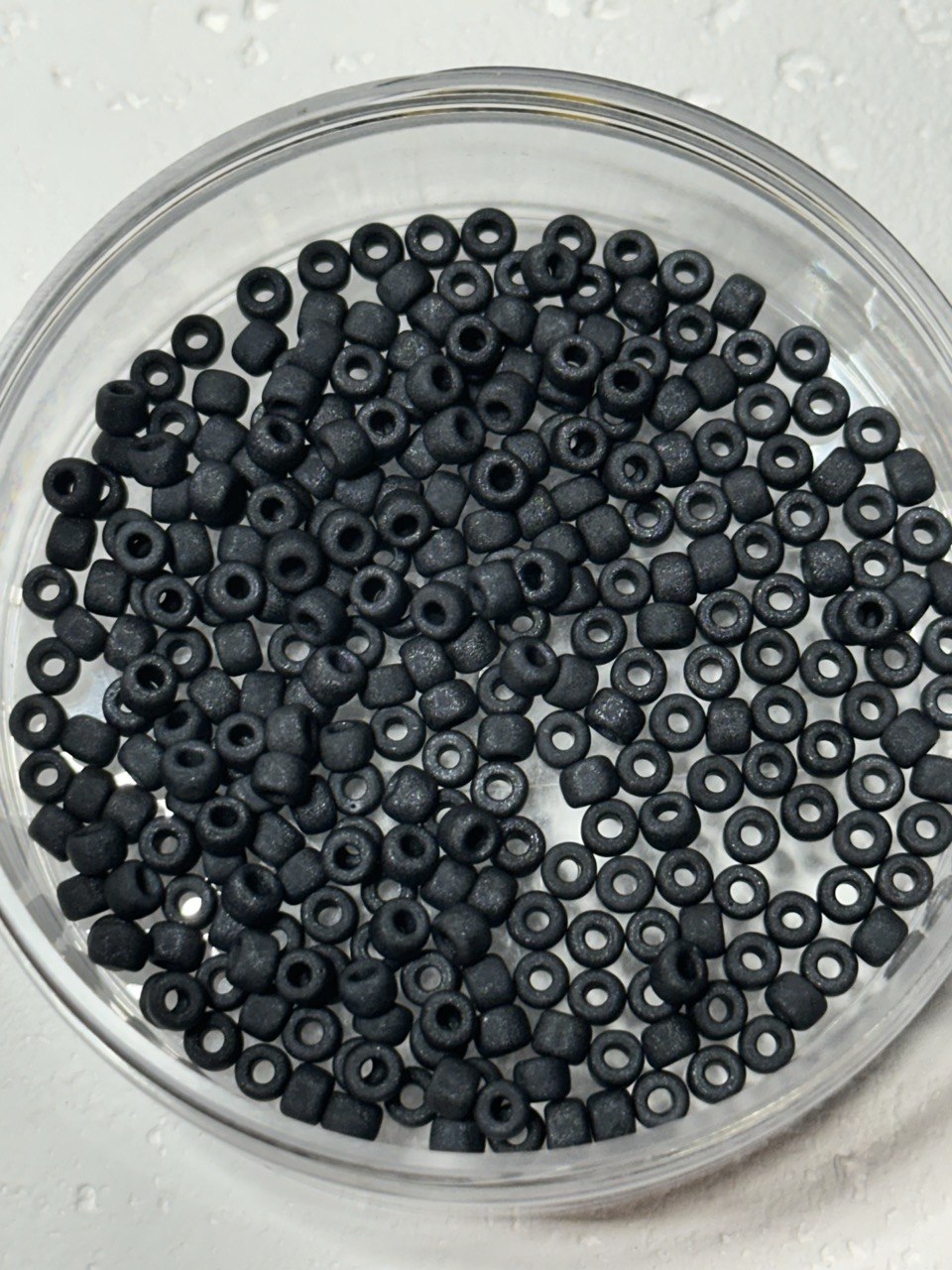 Black seed beads with a matte finish, shown inside a clear plastic bowl on a white textured background.