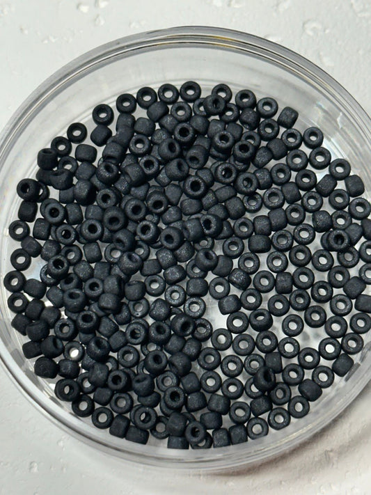 Black seed beads with a matte finish, shown inside a clear plastic bowl on a white textured background.