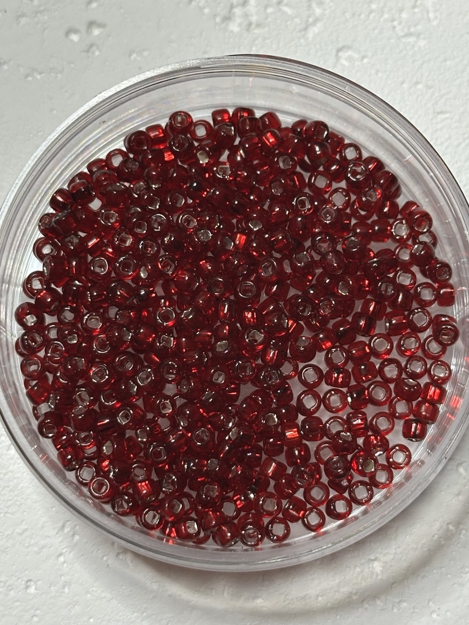 Red glass seed beads piled in a clear round plastic container on a white textured surface.