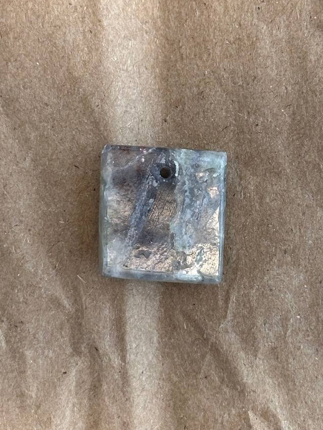 A small square translucent gray and blue stone or glass bead with a top hole, placed on a brown textured paper background.