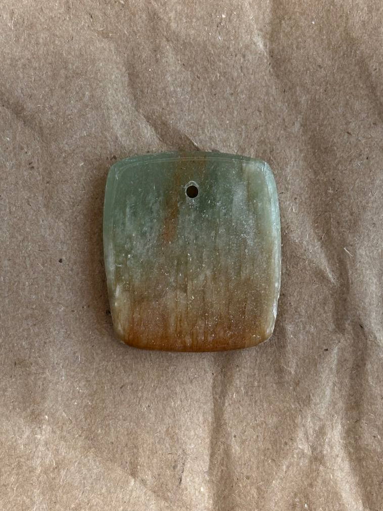 A rectangular stone pendant with rounded corners, featuring a natural color gradient from light green at the top to a warm tan-brown at the bottom, with a small hole near the top center. It is displayed on a textured brown paper background.