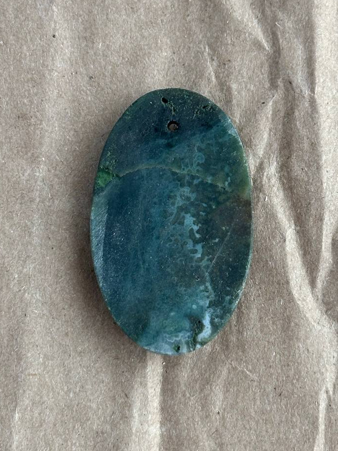 An oval-shaped agate cabochon pendant with a pre-drilled hole, resting on a textured beige paper background.