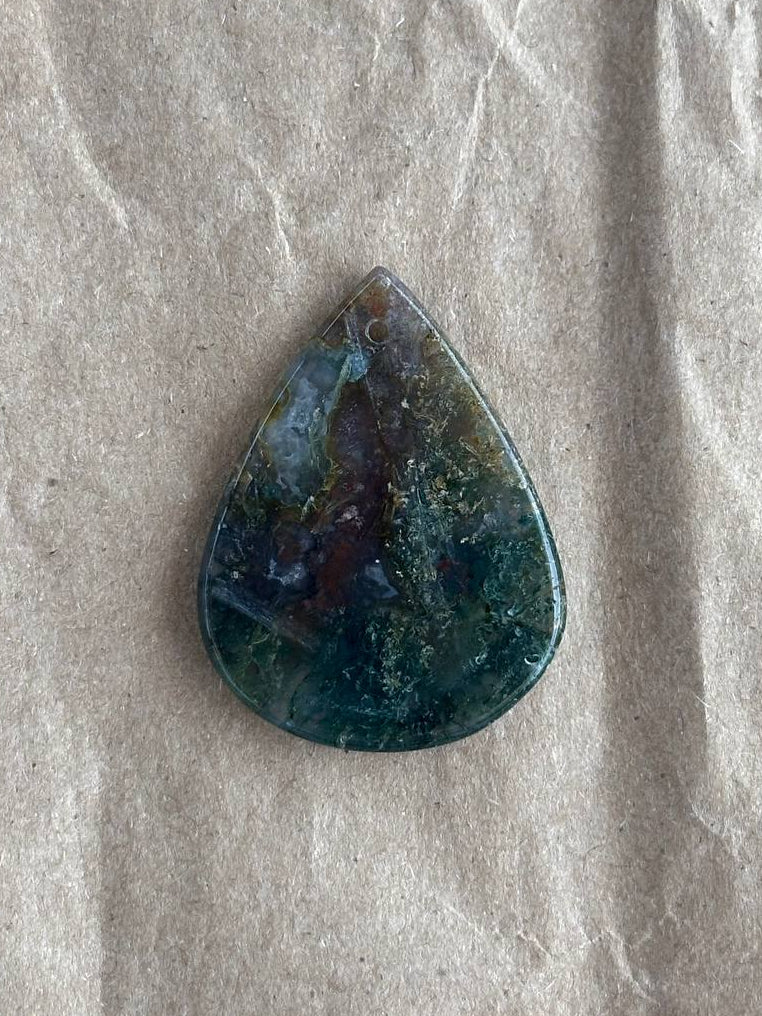 A teardrop-shaped cabochon gemstone with a marbled pattern of dark teal, blue, and brown, resting on a textured beige paper background.