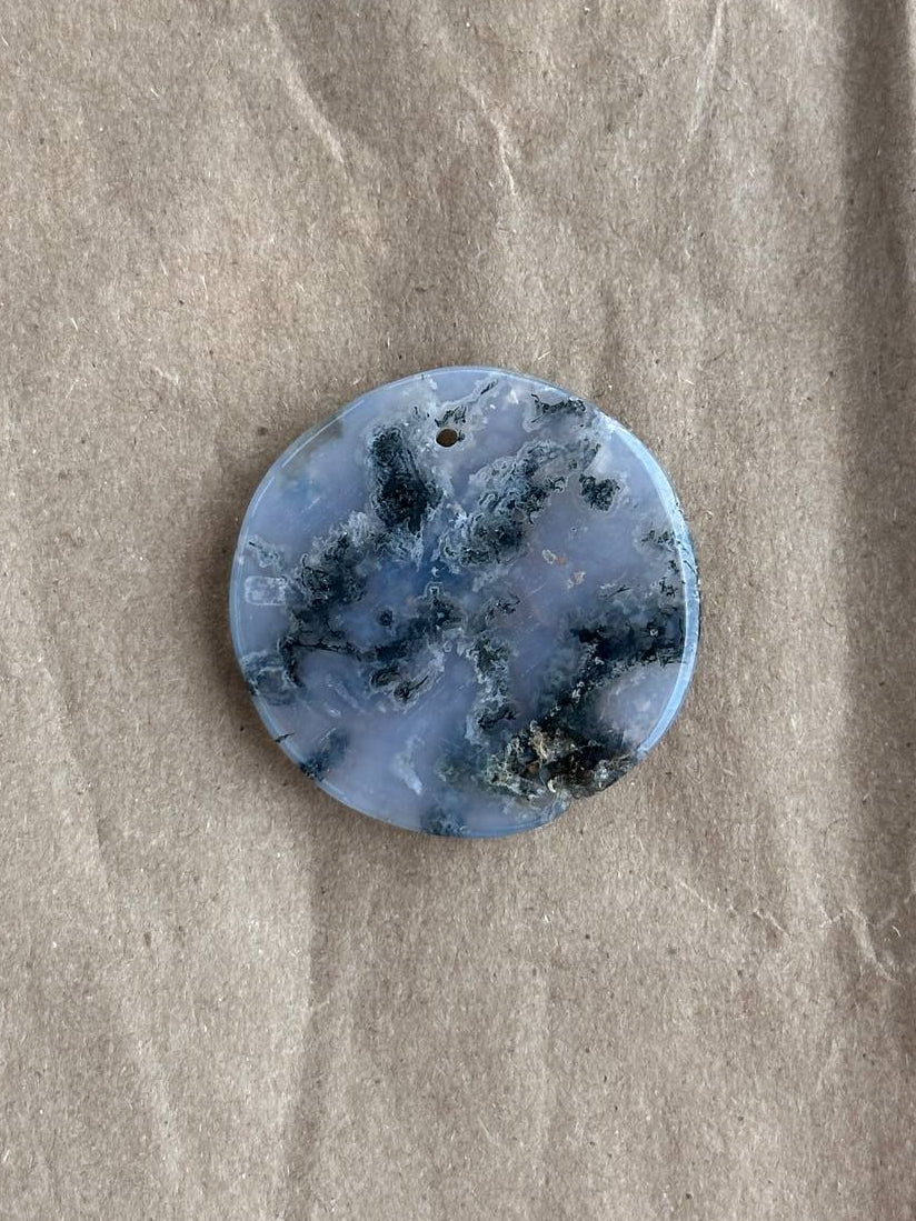 A round, flat blue stone pendant with dark, cloudy inclusions, displayed on a textured beige paper background.