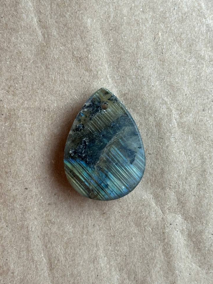 A teardrop-shaped labradorite cabochon gemstone with blue, green, and gold iridescent bands, resting on a textured beige paper background.