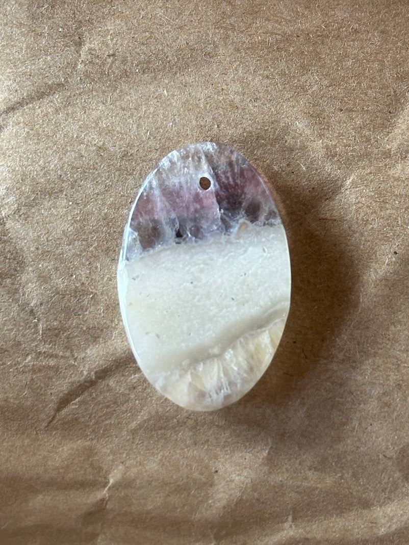An oval-shaped polished stone pendant with a natural marbled pattern of white, gray, and purple, featuring a small hole at the top for hanging, displayed on a brown textured paper background.