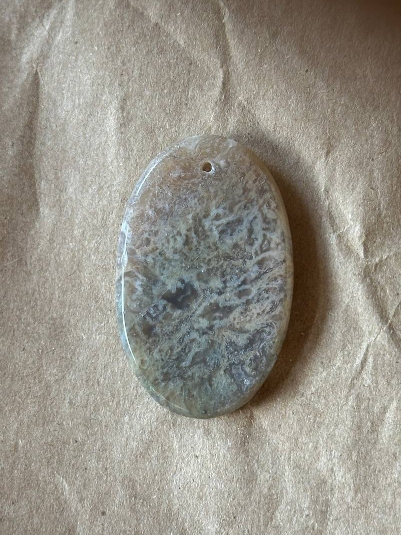An oval stone pendant with a mottled gray and white marbled pattern, featuring a small hole at the top for hanging, displayed on a textured brown paper background.