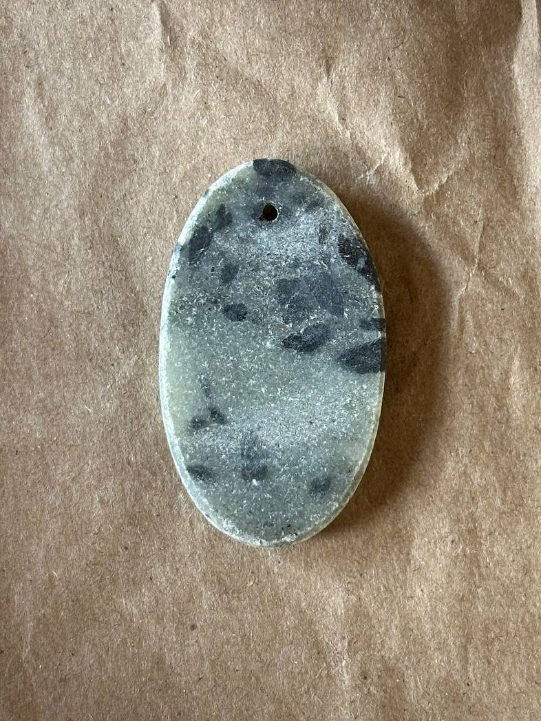 An oval-shaped polished stone pendant with a natural marbled pattern of dark gray and light gray, featuring a small hole at the top for hanging, displayed on a textured brown paper background.