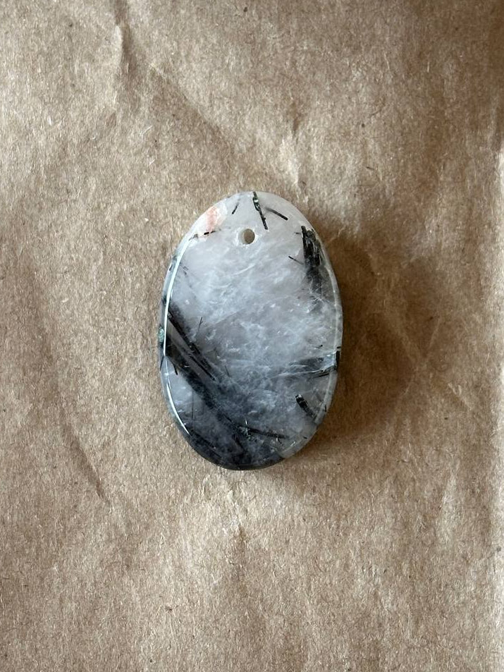 An oval-shaped polished stone pendant with a white, gray, and black marbled pattern, featuring a small hole at the top for hanging, displayed on a textured brown paper background.