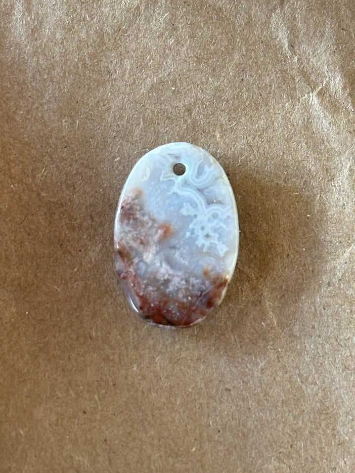 An oval stone pendant with a hole at the top, featuring a light gray and white floral pattern with reddish-brown marbled areas, resting on a textured brown paper background.