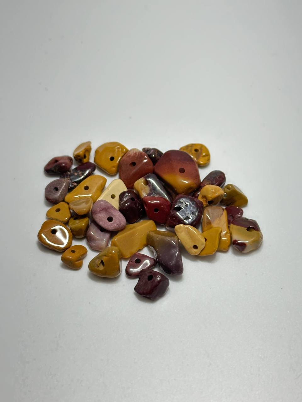 A pile of small, irregularly shaped tumbled gemstone beads in various earthy colors including yellow, brown, red, purple, and pink, each featuring a single hole drilled through the center, set against a plain light gray background.