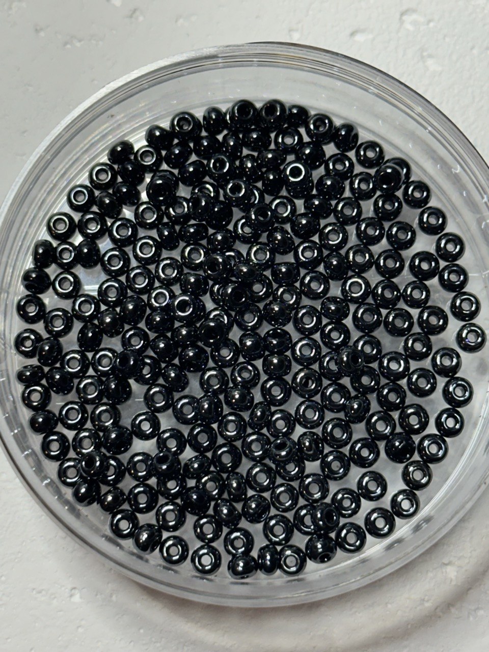 Black seed beads with a glossy finish, shown inside a clear round plastic container on a light textured background.