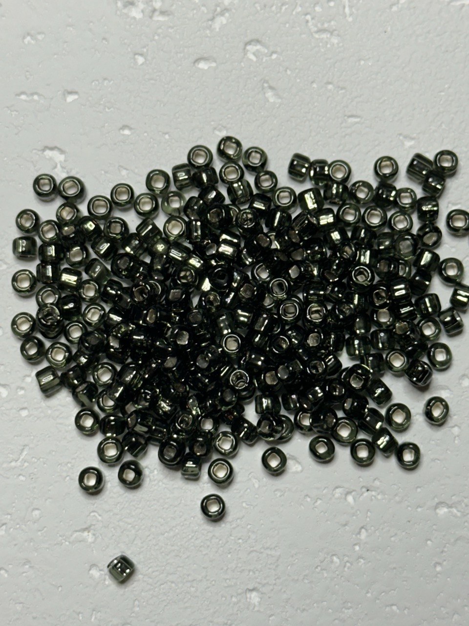 A pile of small metallic dark gray seed beads with a reflective finish, scattered on a light gray textured surface with water droplets.