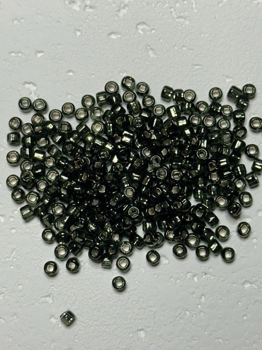 A pile of small metallic dark gray seed beads with a reflective finish, scattered on a light gray textured surface with water droplets.