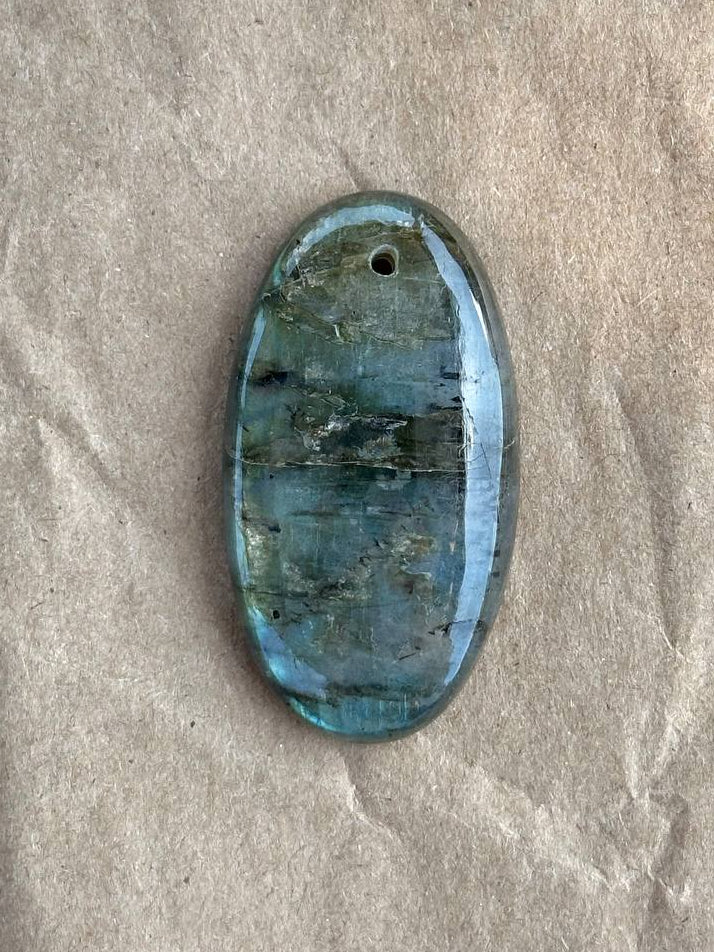 An oval-shaped polished labradorite gemstone pendant with natural blue and gray marbling, displayed on a textured beige paper background.