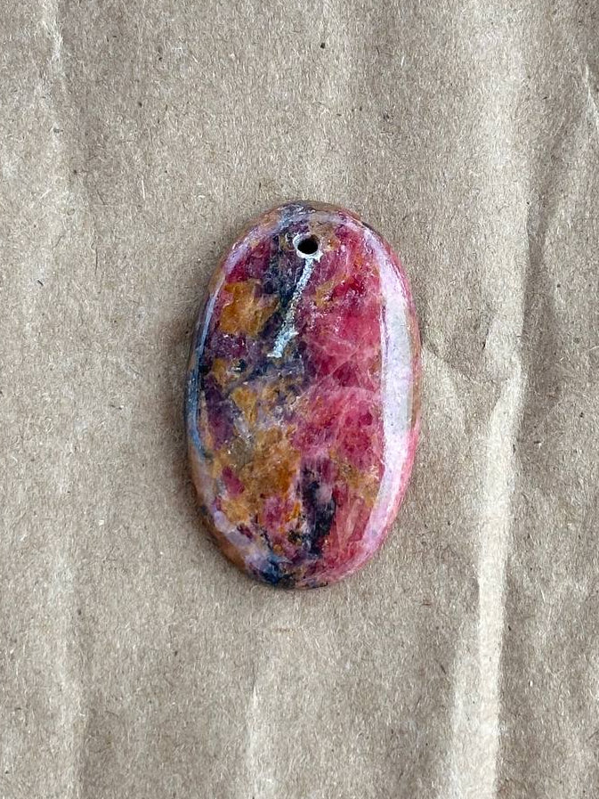 An oval-shaped pendant with a marbled pattern of pink, purple, yellow, and blue, featuring a small hole at the top for stringing, displayed on a textured beige paper background.