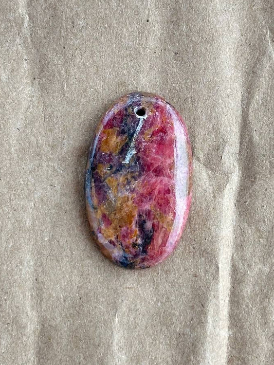 An oval-shaped pendant with a marbled pattern of pink, purple, yellow, and blue, featuring a small hole at the top for stringing, displayed on a textured beige paper background.