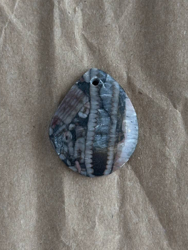 A teardrop-shaped shell pendant with a natural striped pattern in shades of gray, blue, and white, featuring a small hole at the top for hanging, displayed on a textured brown paper background.