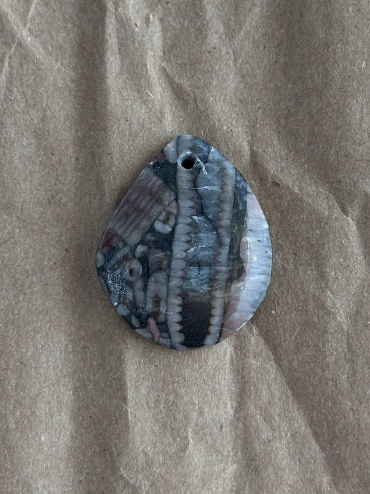 A teardrop-shaped shell pendant with a natural striped pattern in shades of gray, blue, and white, featuring a small hole at the top for hanging, displayed on a textured brown paper background.