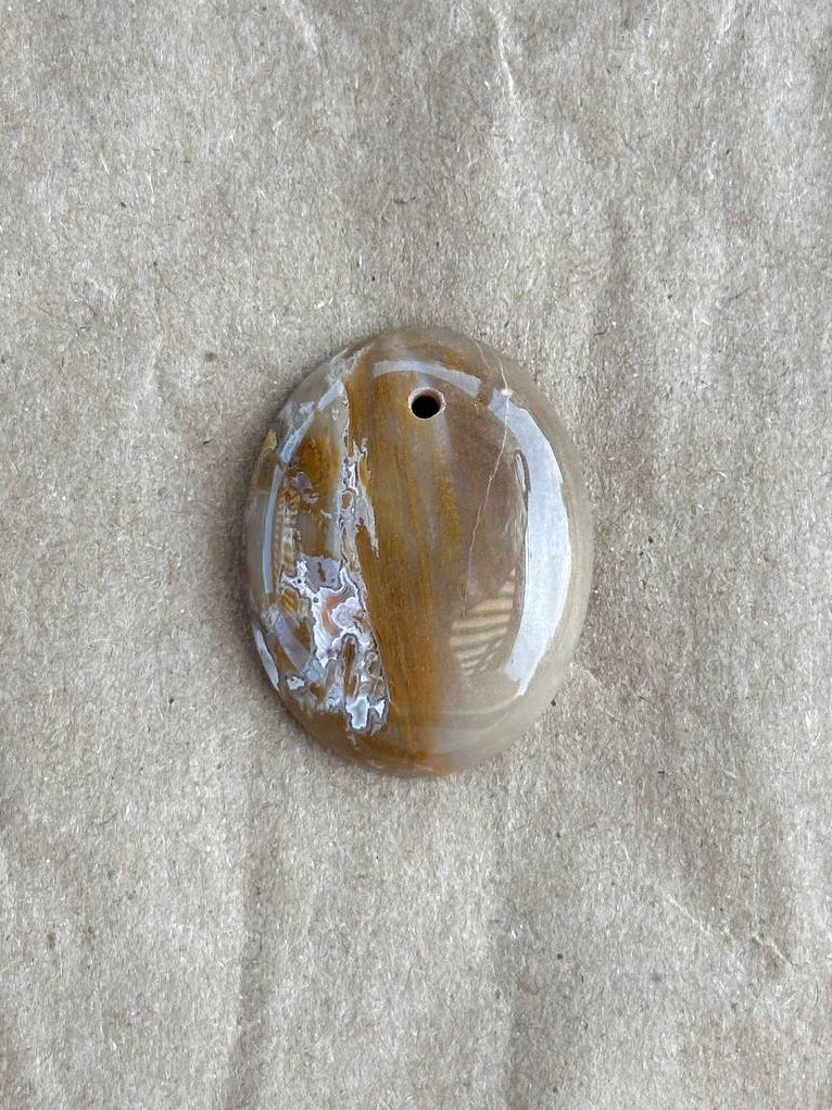 A polished oval agate pendant with a natural banded pattern in brown, beige, and white, featuring a small hole at the top for hanging, displayed on a textured beige paper background.