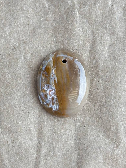A polished oval agate pendant with a natural banded pattern in brown, beige, and white, featuring a small hole at the top for hanging, displayed on a textured beige paper background.