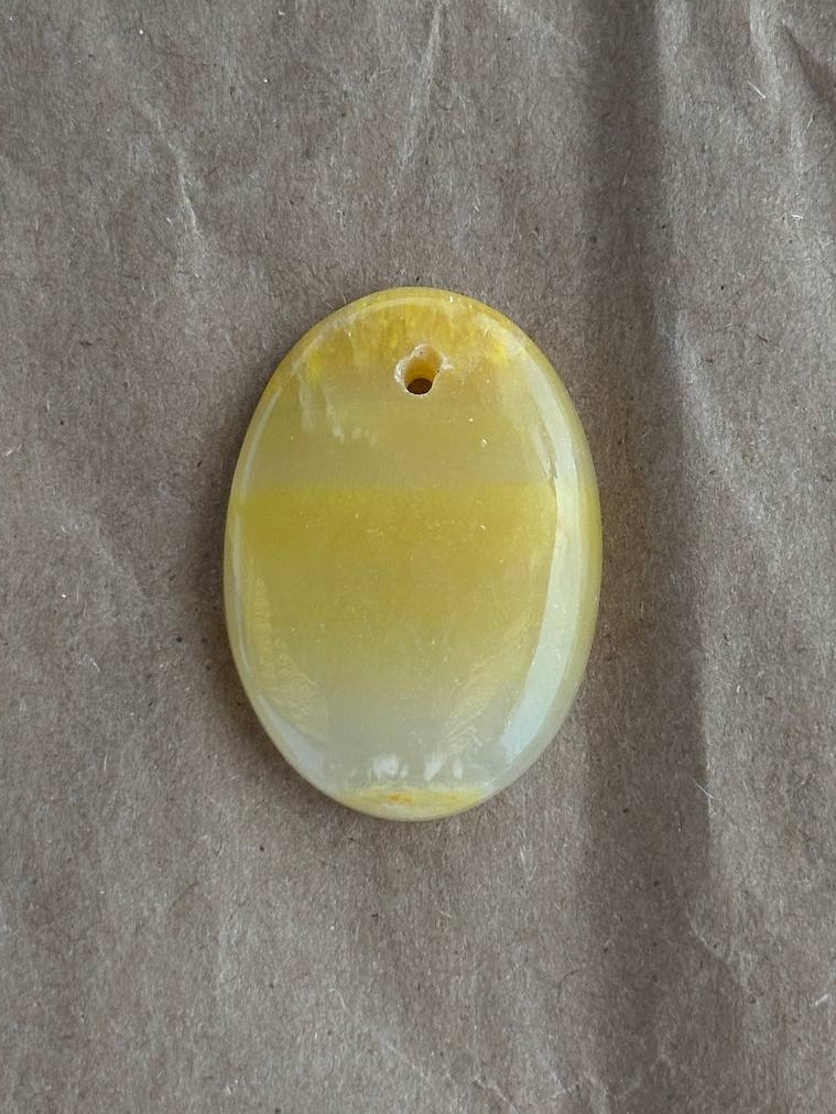 An oval yellow stone pendant with a small hole at the top, resting on a textured gray surface.