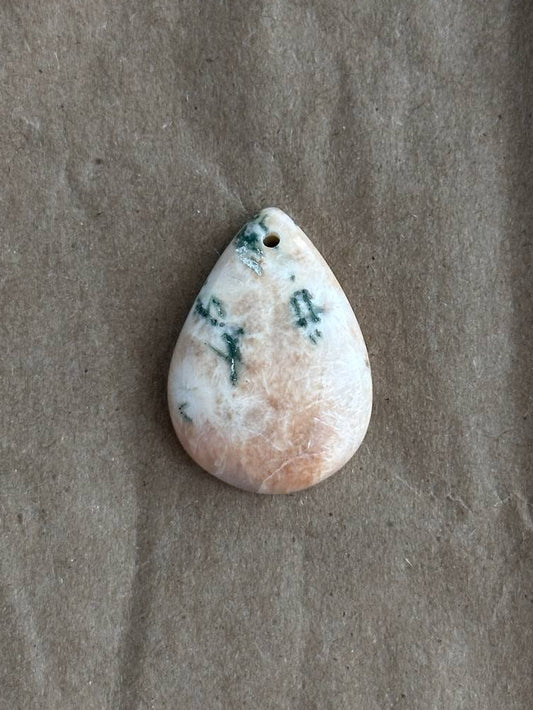 A teardrop-shaped stone pendant with a white and peach-pink marbled pattern and dark green inclusions, displayed on a textured brown paper background.