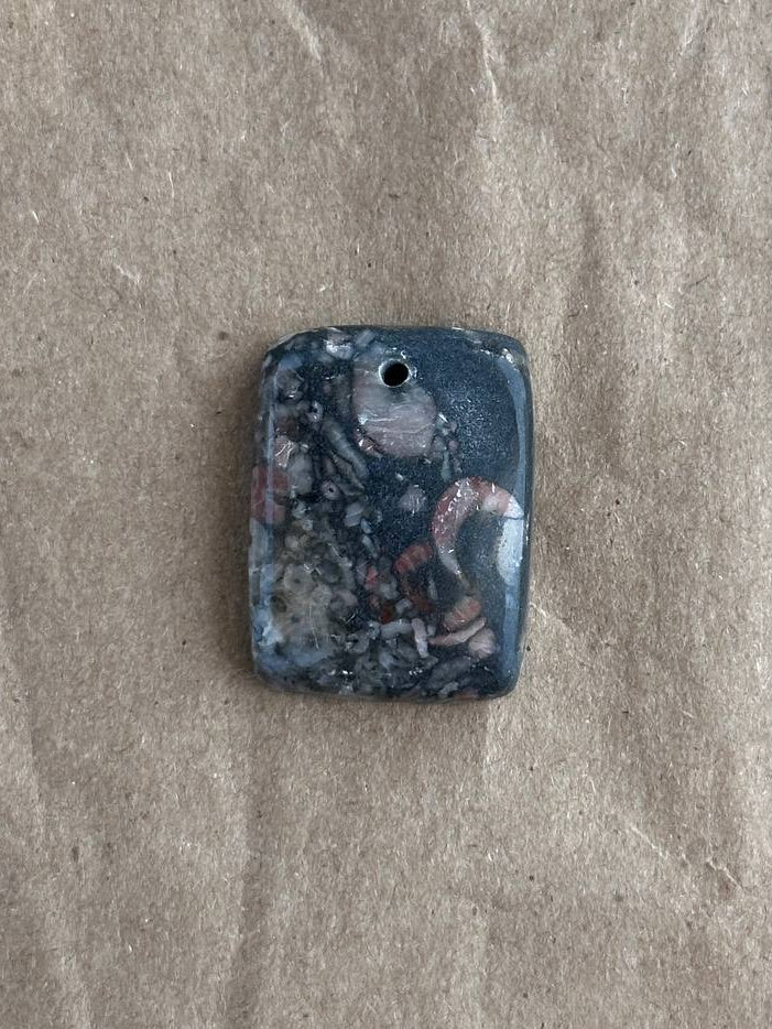 A rectangular dark blue stone pendant with a white and pinkish abstract pattern, featuring a small hole at the top, placed on a textured brown paper background.