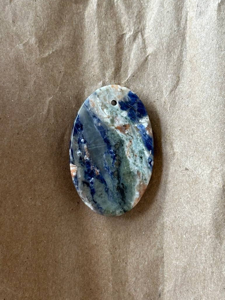 An oval-shaped polished gemstone cabochon with natural blue, white, and gray marbling, featuring a small hole at the top for hanging, displayed on a textured brown paper background.