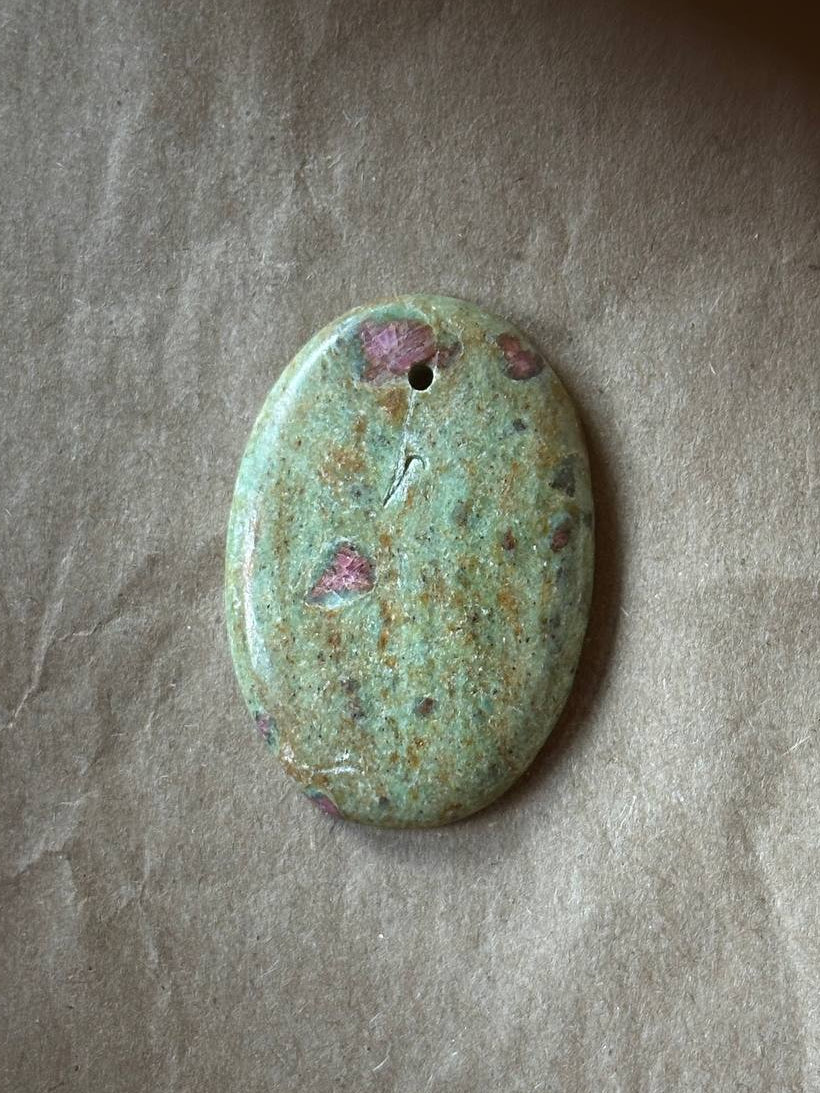 An oval polished stone pendant with a natural speckled pattern of green, beige, and pinkish-purple, featuring a small hole at the top for hanging, displayed on a textured brown paper background.
