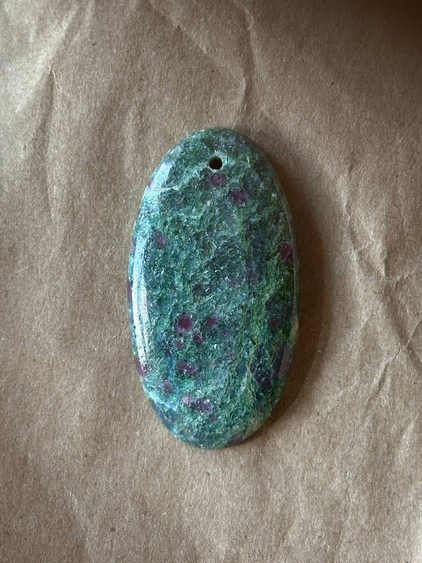 An oval-shaped polished gemstone pendant with a green and blue marbled pattern and purple inclusions, featuring a small hole at the top, displayed on a textured brown paper background.
