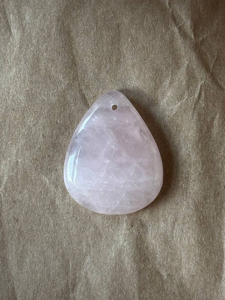 A teardrop-shaped pale pink stone pendant with a small hole at the top, resting on a textured brown paper background.