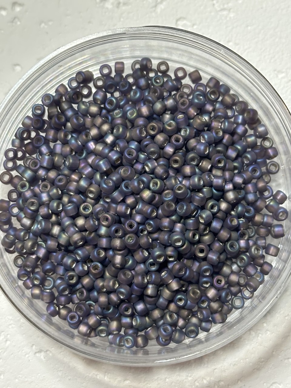 A top-down view of small iridescent seed beads in a clear round plastic container, showing a metallic purple and blue shimmer on a white textured background.