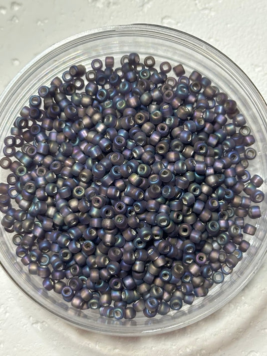 A top-down view of small iridescent seed beads in a clear round plastic container, showing a metallic purple and blue shimmer on a white textured background.