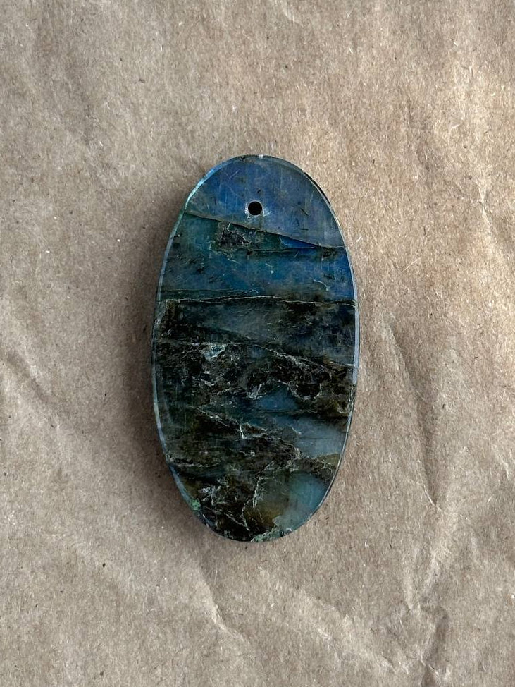 An oval blue labradorite stone pendant with natural dark veining and a small hole at the top, displayed on a textured beige paper background.