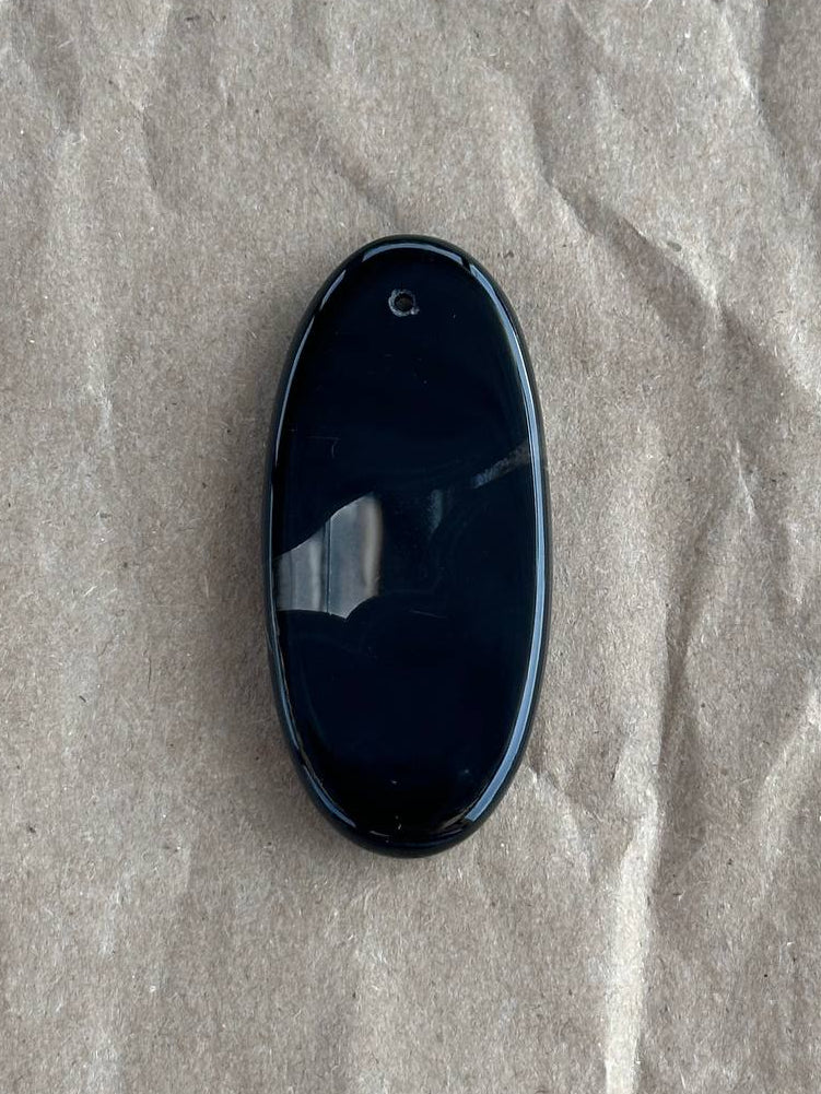 A glossy black oval pendant or charm with a small loop at the top, resting on a textured, crumpled light brown paper background.