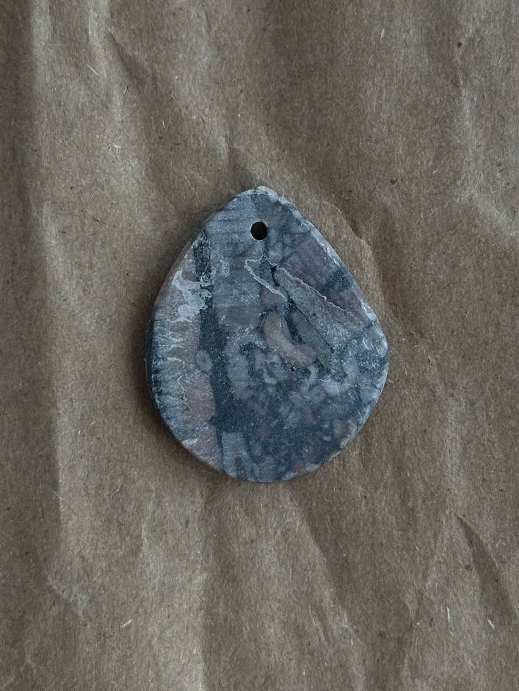 A teardrop-shaped stone pendant with a mottled blue, gray, and white pattern, featuring a small hole at the top for hanging, displayed on a textured brown paper background.