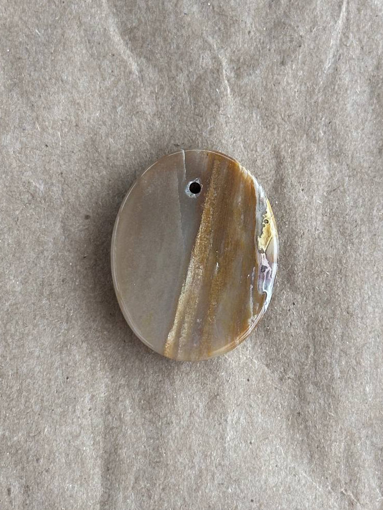 An oval agate pendant with a natural striped pattern in beige, brown, and cream, featuring a small hole at the top for hanging, displayed on a textured gray fabric background.