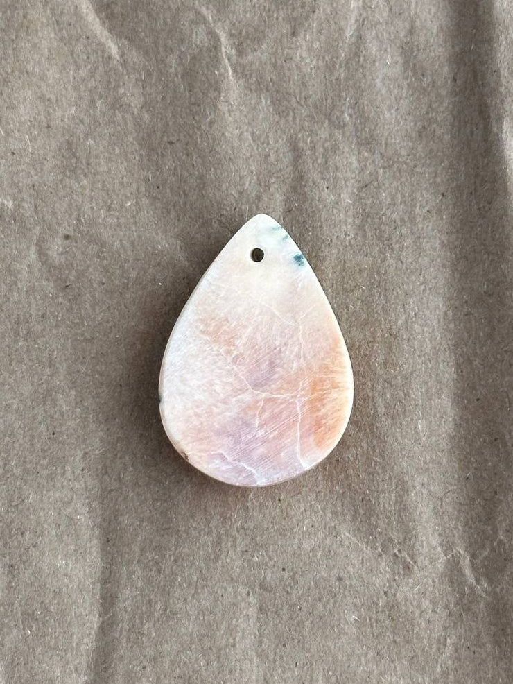 A teardrop-shaped pendant made from a polished natural stone with pink, peach, and white marbling, featuring a small hole at the top for hanging. The pendant is displayed on a textured, wrinkled brown paper background.