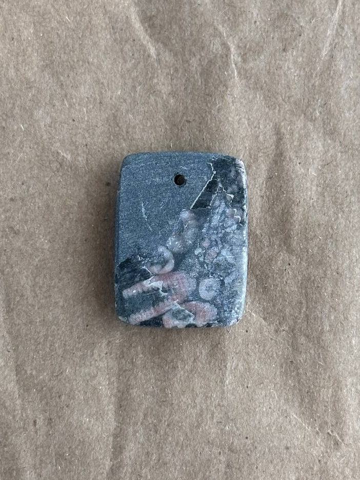 A rectangular stone pendant with rounded corners, featuring a marbled pattern of blue, gray, and white, complete with a small hole at the top for hanging, placed on a textured brown paper background.